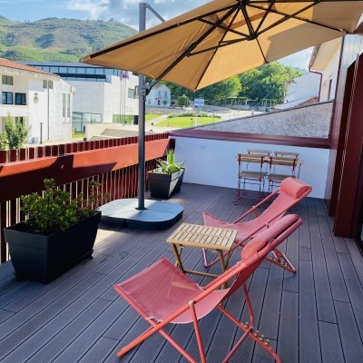 Varanda com espreguiçadeiras coral, guarda-sol bege, floreiras pretas e mesa com cadeiras de madeira