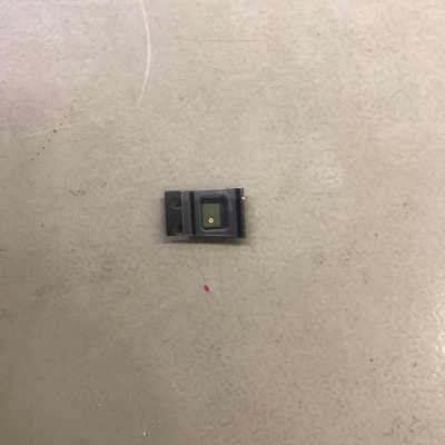 Componente eletrônico pequeno cinza escuro com detalhe amarelo no centro sobre superfície bege