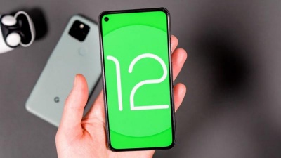 Smartphone cinza claro com ecrã verde e número 12 mostrado na mão, outro igual e auscultadores sem fios ao fundo