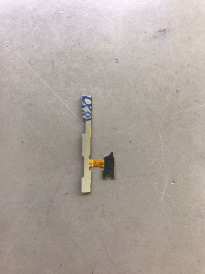 Componente eletrónico pequeno com fita flexível amarela e partes metálicas cinzentas sobre superfície riscada