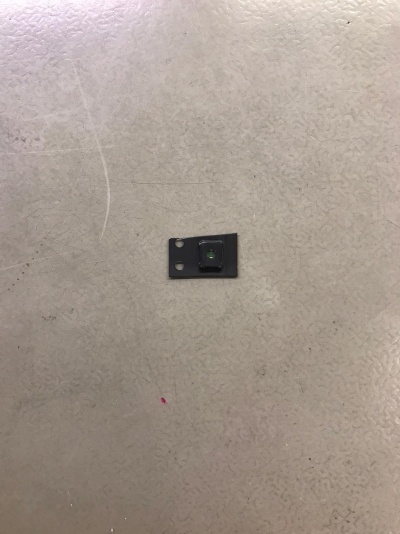 Componente eletrónico preto quadrado com lente central sobre superfície cinzenta