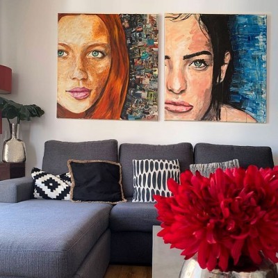 Sofá cinzento com almofadas, duas pinturas de rostos femininos, vaso com planta e flores vermelhas.