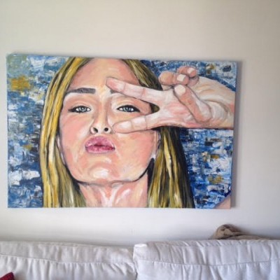 Tela pintada retratando mulher loira a fazer sinal de paz, pendurada em parede branca acima de sofá cinza claro