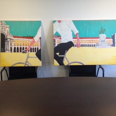 Dois quadros pintados com uma pessoa a andar de bicicleta numa praça com edifícios e estátua