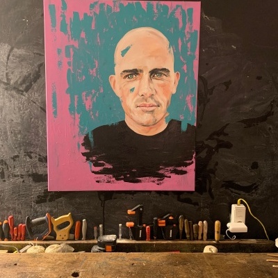 Pintura de retrato moderno de homem careca com camisa preta em parede preta acima de bancada de madeira e ferramentas.