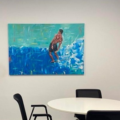 Quadro colorido com pessoa em tronco nu sobre fundo azul e verde, pendurado em parede branca acima de mesa redonda e cadeiras pretas