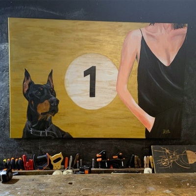 Quadro com cão Doberman, número 1 e mulher com top preto numa parede escura, bancada de madeira com ferramentas em frente.