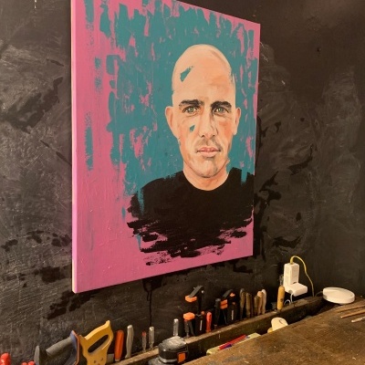 Tela pintada com retrato de homem calvo com fundo rosa e azul, acima de bancada com ferramentas