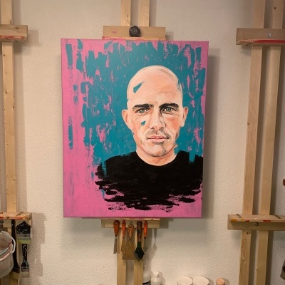 Quadro pintado de rosto de homem careca com fundo azul e rosa em cavalete de madeira.