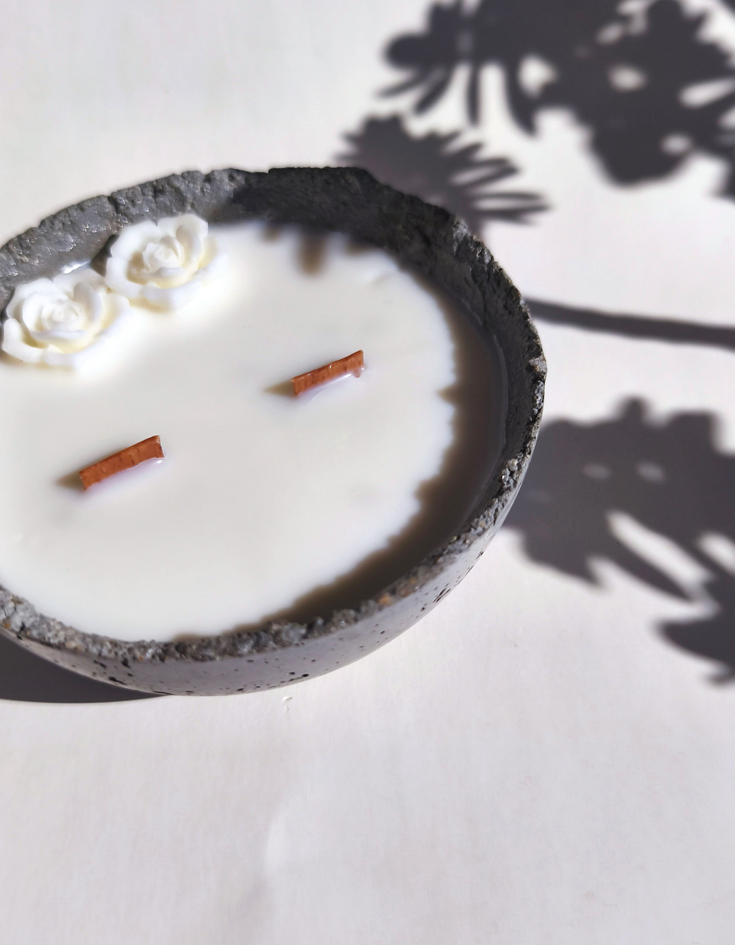 Vela Aromática Bowl - Bergamota e Sândalo Vela artesanal branca numa base cinza escura com flores em relevo e sombra de folhas no fundo claro