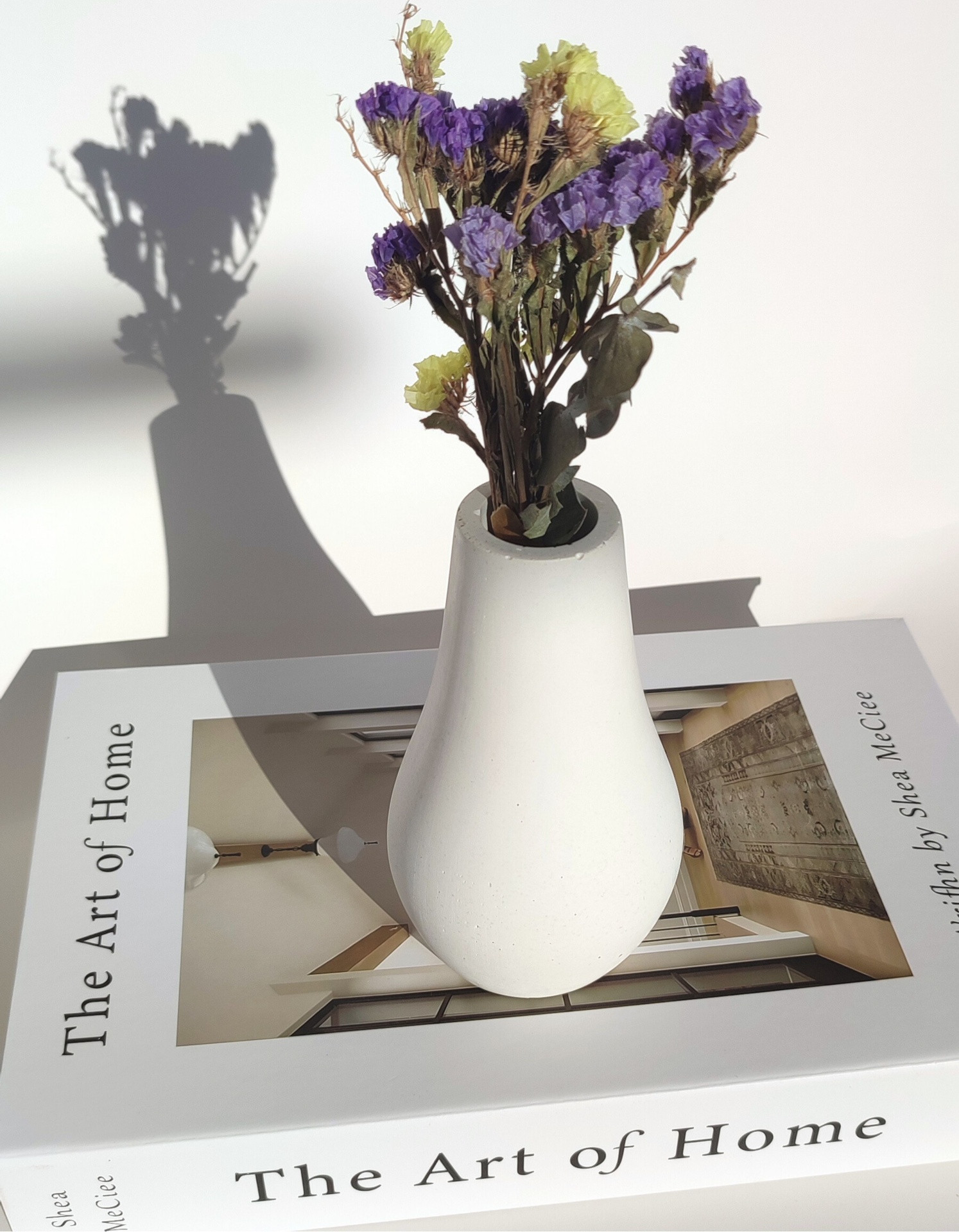 Jarra Lotus Minimal Vaso branco com flores secas sobre livro 'The Art of Home'