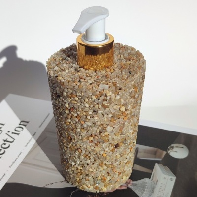 Dispenser cilíndrico revestido por pedras naturais com tampa branca e anel dourado sobre revista