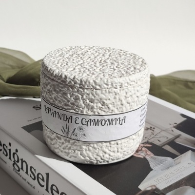 Frasco redondo branco texturado com rótulo AMANDA E CAMOMILA sobre livro e pano verde
