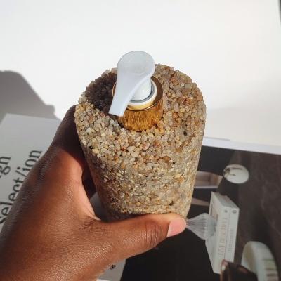 Frasco dispensador com exterior de pedras, tampa branca e válvula dourada, segurado por mão, fundo com livro e foto.