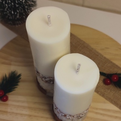 Duas velas brancas cilíndricas com textura granulada na base, sobre base de madeira clara, com ramos e bagas vermelhas decorativas ao lado