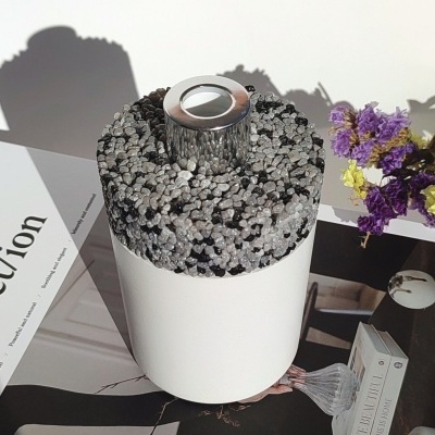 Vela decorativa branca com topo em pedras pretas e brancas, tampa prateada, sobre revista e flores secas