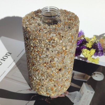 Garrafa cilíndrica revestida com pequenas pedras coloridas em cima de revista com flores secas ao fundo