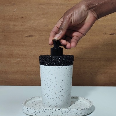 Dispenser de sabão líquido branco e preto com textura granulada numa base oval, com mão a premir o bico