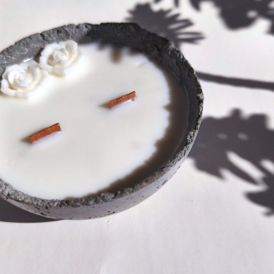 Vela artesanal branca numa base cinza escura com flores em relevo e sombra de folhas no fundo claro