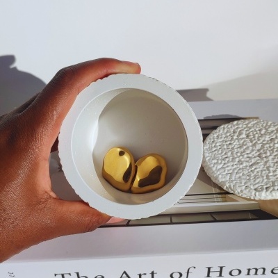 Recipiente branco com dois brincos dourados e livro ao fundo