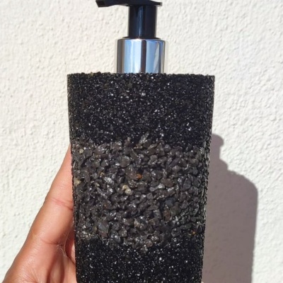 Dispenser de bomba para líquido com base texturizada preta e pedras negras, tampa prateada, segurado por mão.