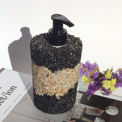 Doseador de líquido com textura de pedras em preto e bege, bomba preta, ao lado de flores e livros.