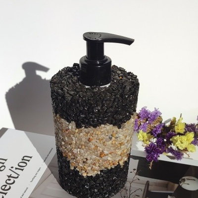 Frasco dispensador texturizado com pedras e tampa preta ao lado de flores secas e revista aberta.