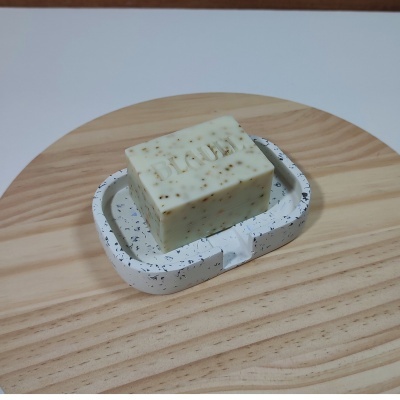 Sabonete branco com partículas castanhas numa saboneteira de cerâmica branca com manchas pretas sobre madeira clara