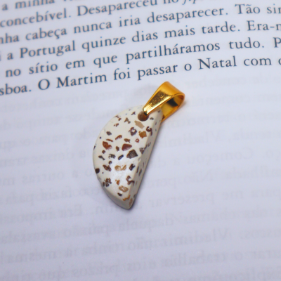 Pendente branco com manchas castanhas e argola dourada sobre texto em papel.