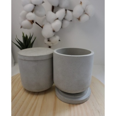 Duas caixas cinzentas de cimento, uma com tampa e outra aberta, em superfície de madeira com ramo de algodão e planta ao fundo