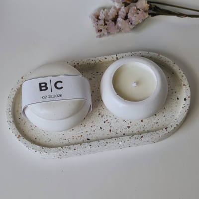 Conjunto de vela com base e tampa em cerâmica branca, sobre suporte retangular manchado, com flores secas ao fundo.