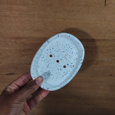 Saboneteira oval branca com pontilhado preto e três furos, segurada sobre fundo de madeira.