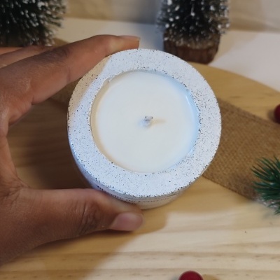 Vela branca em castiçal texturizado branco sendo segurada por uma mão, com árvores decorativas ao fundo