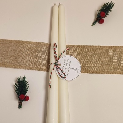 Duas velas brancas amarradas com cordão tricolor e etiqueta redonda, sobre fundo bege com dois ramos verdes com bagas vermelhas.
