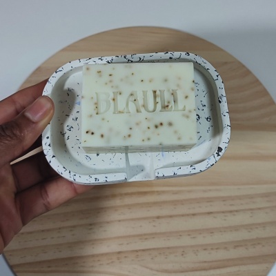 Barra de sabão branca com partículas e saboneteira branca com padrão preto segurada por uma mão