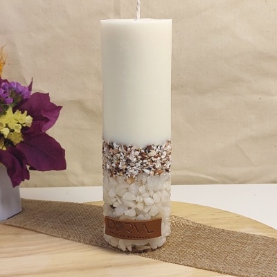 Vela branca decorativa com pedras naturais e etiqueta DORVAL sobre mesa de madeira com flores ao lado.