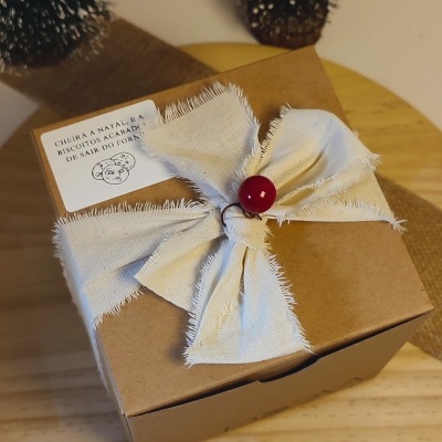 Caixa de cartão castanha com laço branco e bola vermelha, etiqueta com texto, sobre mesa de madeira.