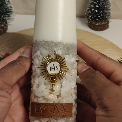 Vela branca alta com base decorada com pedras e areia, selo dourado IHS e etiqueta de couro DORAL arte em velas