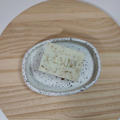 Barra de sabão branca com partículas e texto BLAUBL na saboneteira oval cinza com pontos pretos sobre madeira clara