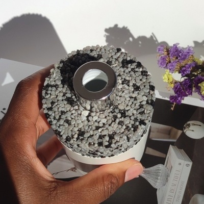 Frasco branco com tampa de pedras cinza e preto segurado por mão, flores roxas e amarelas ao lado