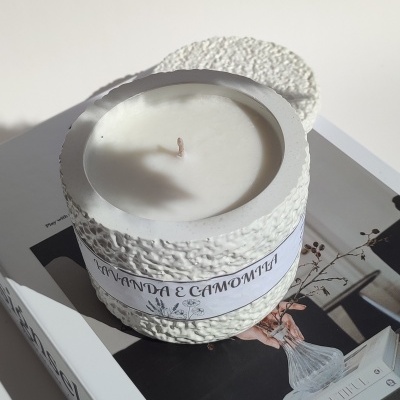 Vela branca em jarra de cerâmica texturizada colocada sobre um livro com imagem de mesa e vase de flores.