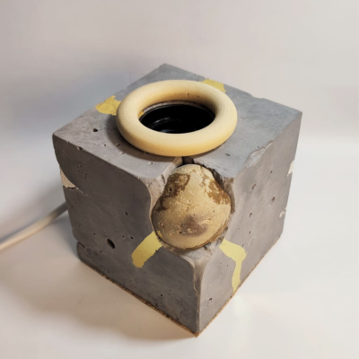 Luminária cúbica de cimento com suporte circular bege e detalhes amarelos, sobre fundo branco.