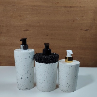 Três dispensadores de sabonete líquidos cerâmicos branco com manchas pretas e tampas de plástico, sobre superfície branca e fundo de madeira clara