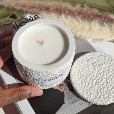 Vela branca em recipiente texturizado branco com tampa e etiqueta, sobre revista e decorada com ervas secas e tecido verde