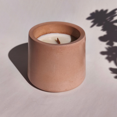 Vela branca num recipiente cilíndrico rosa com sombra de planta ao fundo