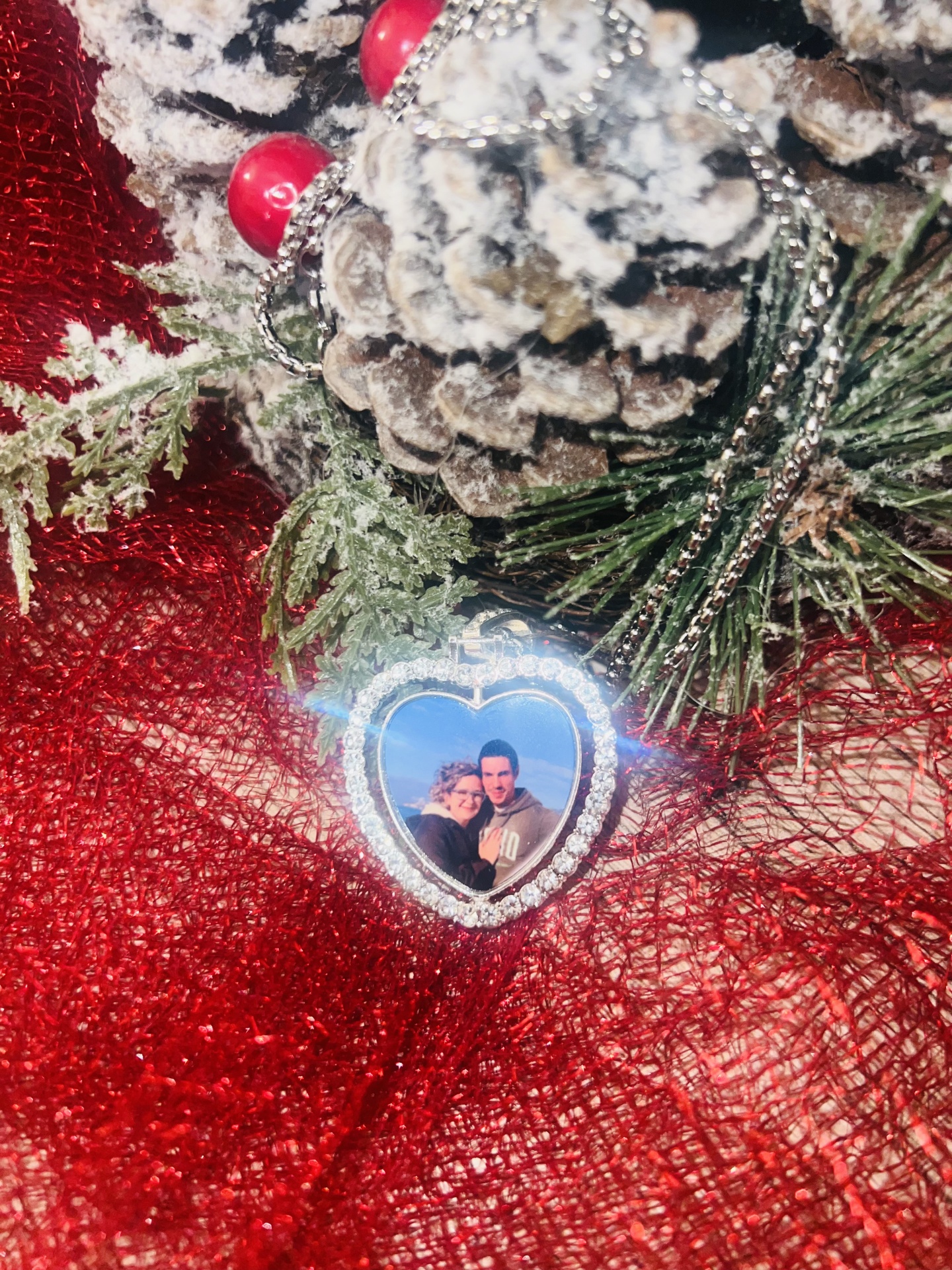 Colar com medalhão em forma de coração com fotografia, decorado com brilhantes, sobre tecido vermelho com pinhas e folhas decorativas nevadas.