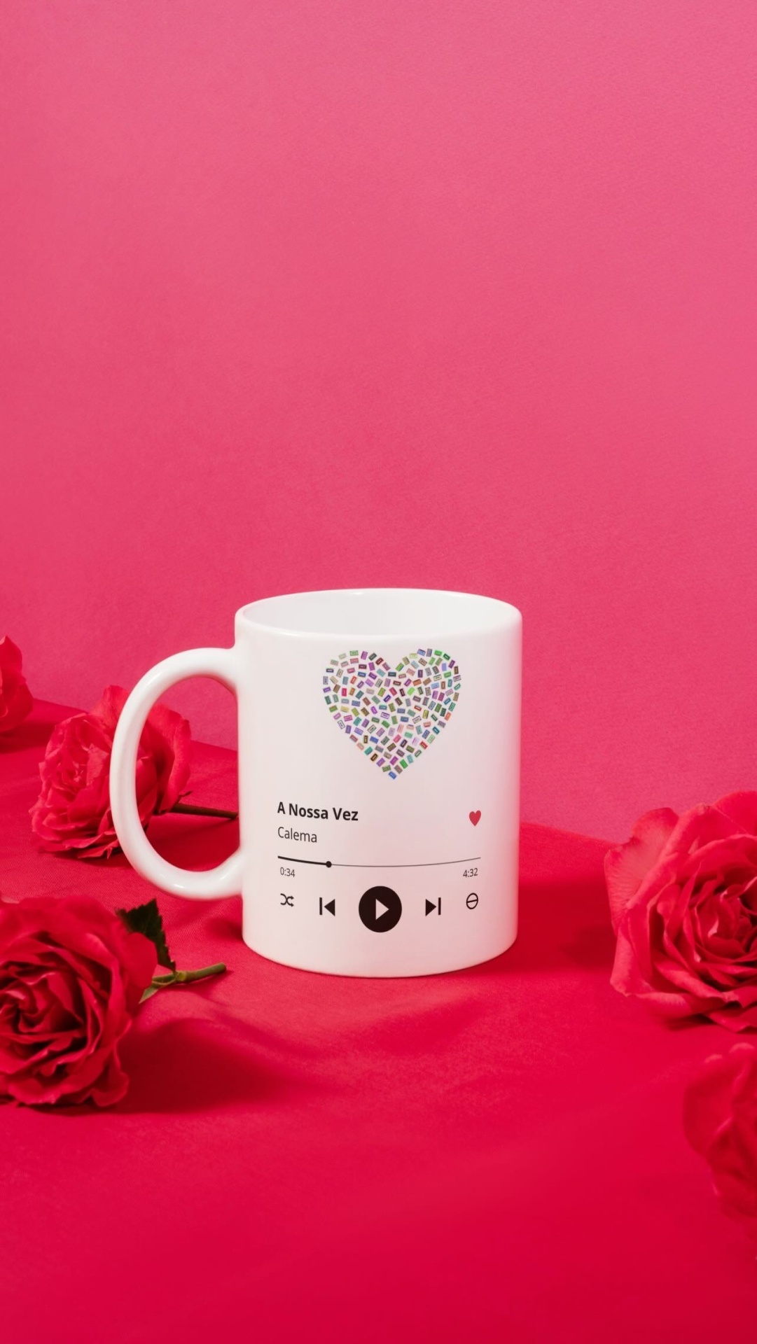 Caneca branca decorada com coração colorido e texto de música numa superfície vermelha com rosas vermelhas.