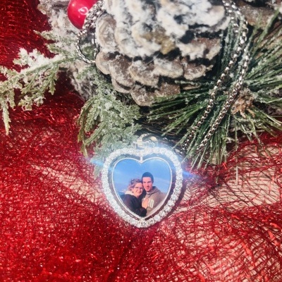 Colar com medalhão em forma de coração com fotografia, decorado com brilhantes, sobre tecido vermelho com pinhas e folhas decorativas nevadas.