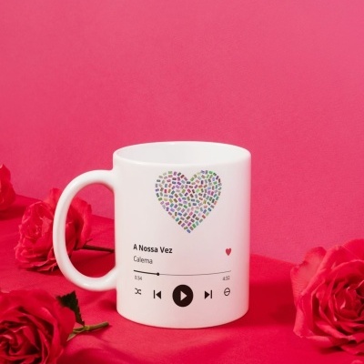Caneca branca decorada com coração colorido e texto de música numa superfície vermelha com rosas vermelhas.