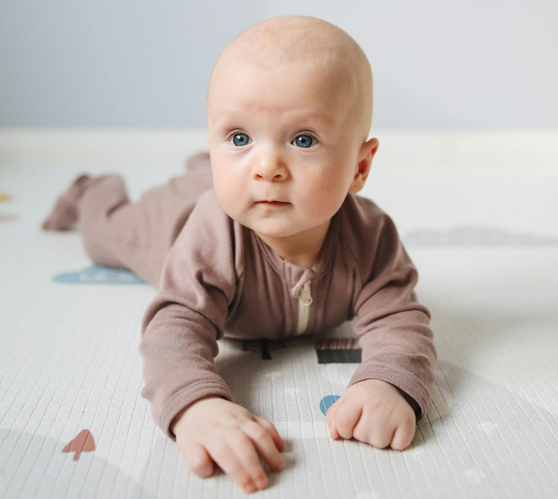Bebé deitado de barriga para baixo com fato castanho claro sobre tapete branco com padrões coloridos